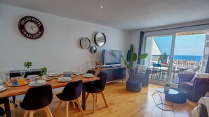 Un salon moderne avec un coin repas, de grandes fenêtres offrant une vue sur la mer et une enseigne du Grand Hotel Charleston accrochée au mur.