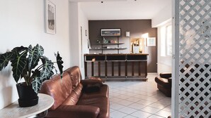 Una sala moderna con un sofá de cuero marrón, una mesa redonda y una planta grande en maceta. Se ve una cocina con barra y estanterías al fondo.