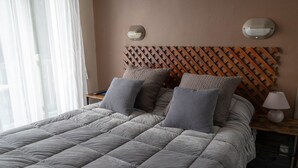 Una cama impecablemente hecha con colcha gris y almohadas estampadas, cabecera de entramado de madera y lámparas de noche.