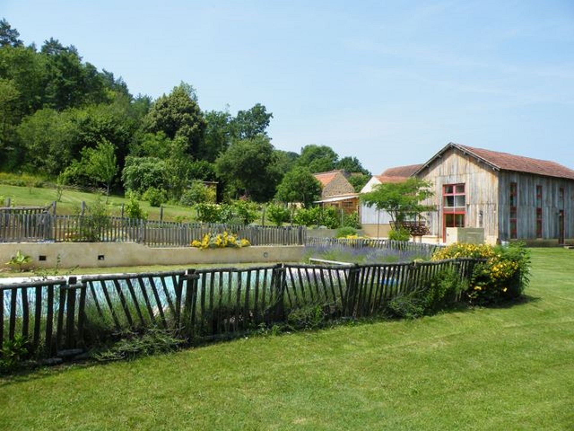 A rustic barn with a red roof, surrounded by a fenced garden with various plants and a small pond.
