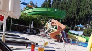Un área junto a la piscina con sillas reclinables, una mesa con bebidas y fruta, y un tobogán al fondo.