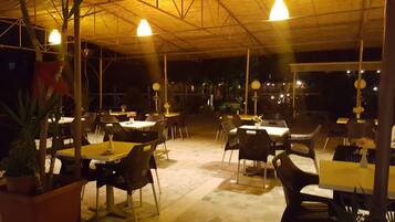Restaurante ao ar livre com mesas e cadeiras, sob uma área coberta com luminárias pendentes.