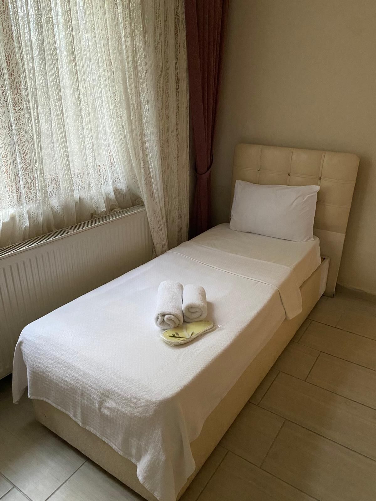 Una cama individual con una colcha blanca, dos toallas enrolladas y una almohada blanca.