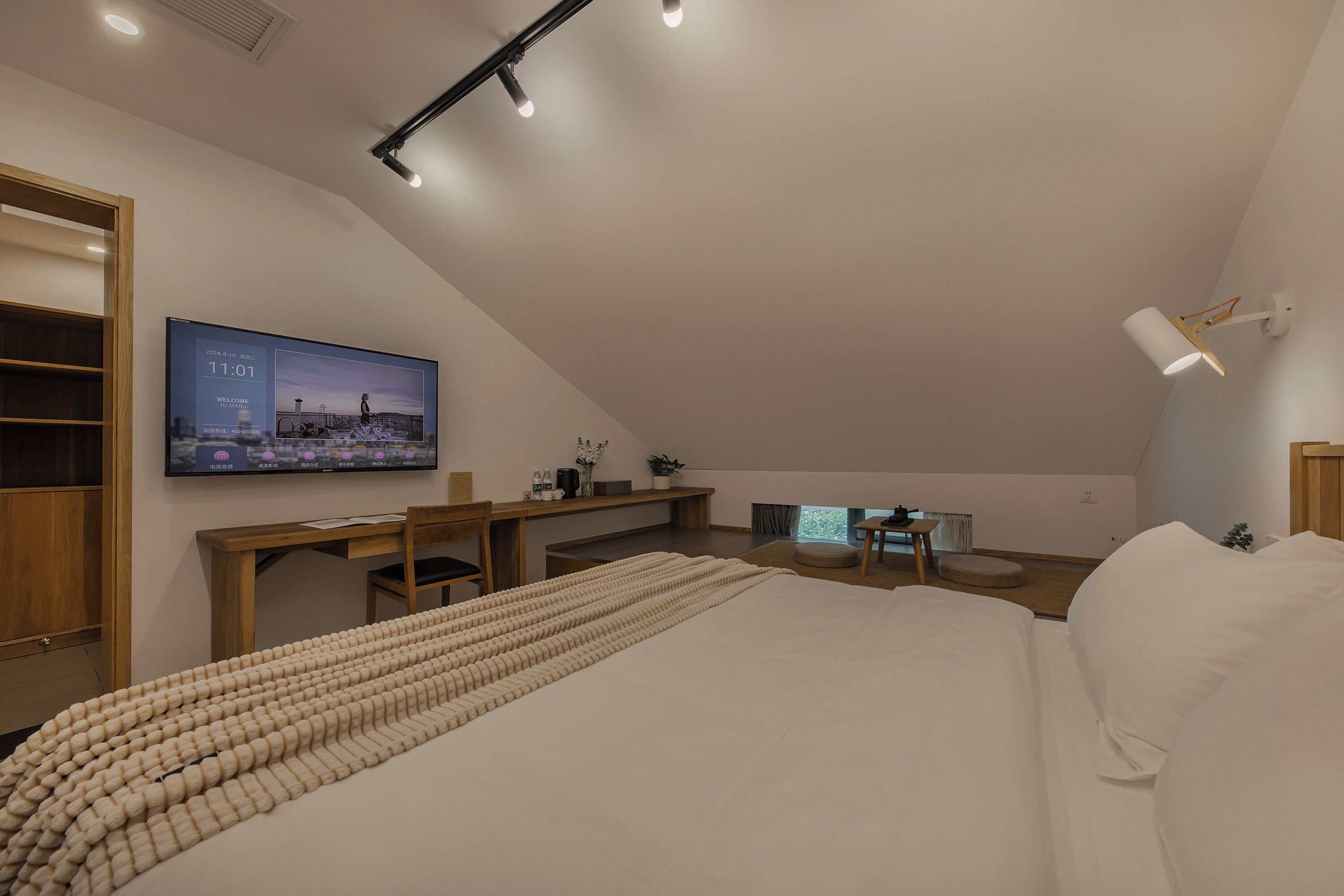 A modern bedroom with a sloped ceiling, a large bed, a desk, and a television displaying a welcome message.