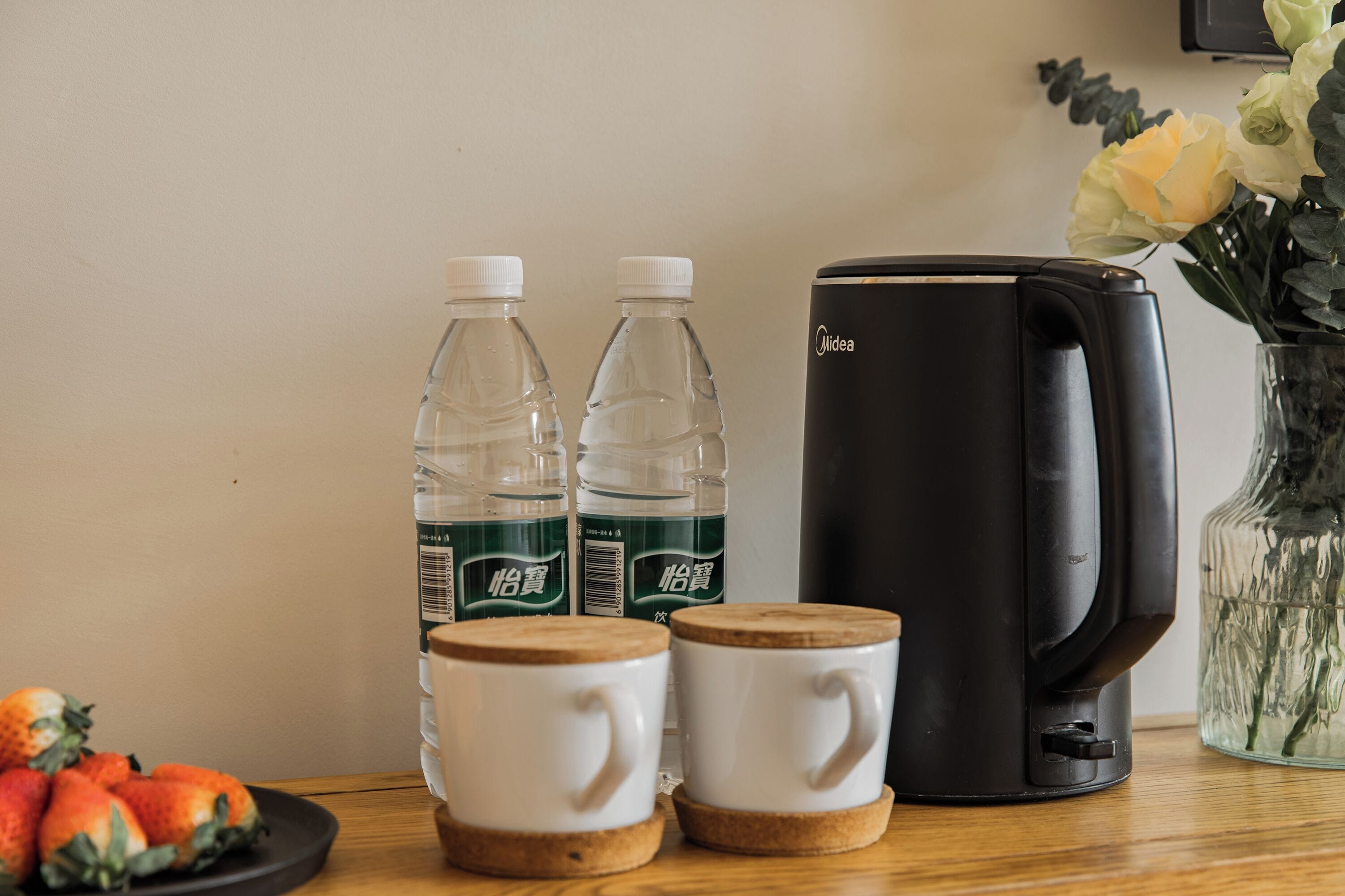 A coffee maker, two water bottles, two white mugs, a plate of strawberries, and a vase with flowers on a wooden table.