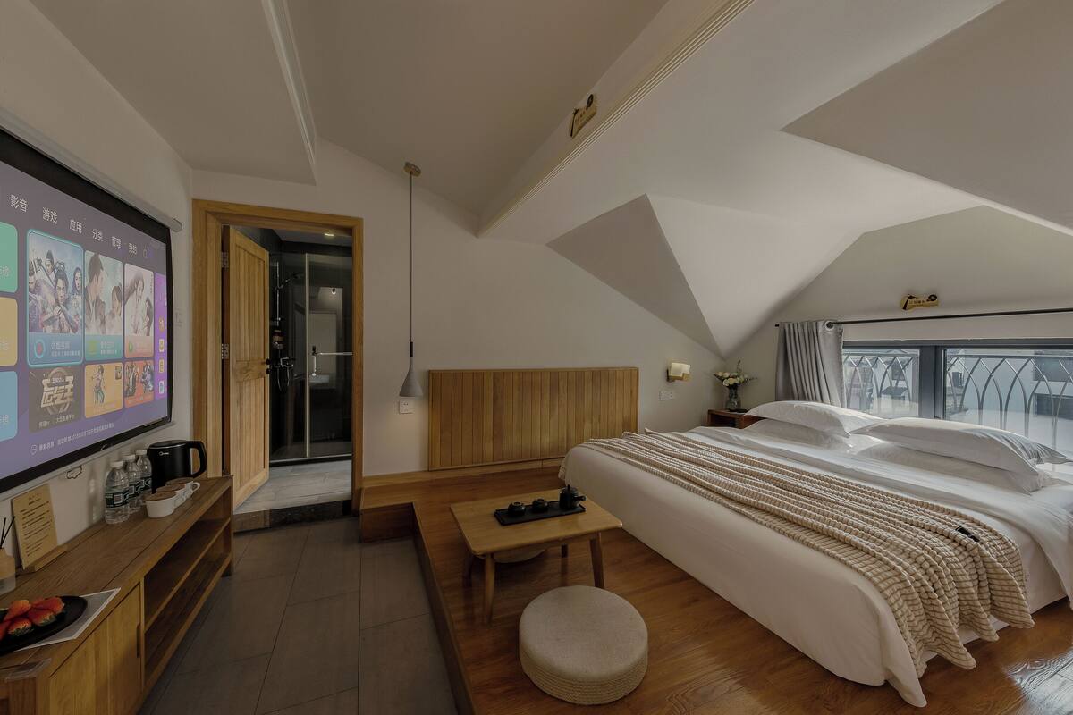 Ein modernes Hotelzimmer mit einem großen Bett, einem hölzernen Kopfteil, einem an der Wand montierten Fernseher, einem kleinen Tisch und einem runden Hocker.