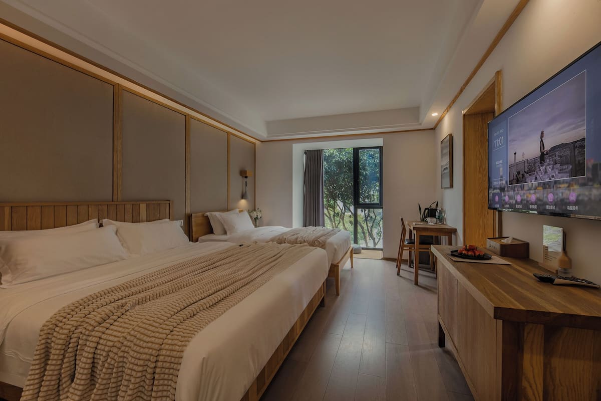 Ein Hotelzimmer mit zwei Betten, einem Holztisch, einem Fernseher und einem Balkon mit Blick auf Bäume.