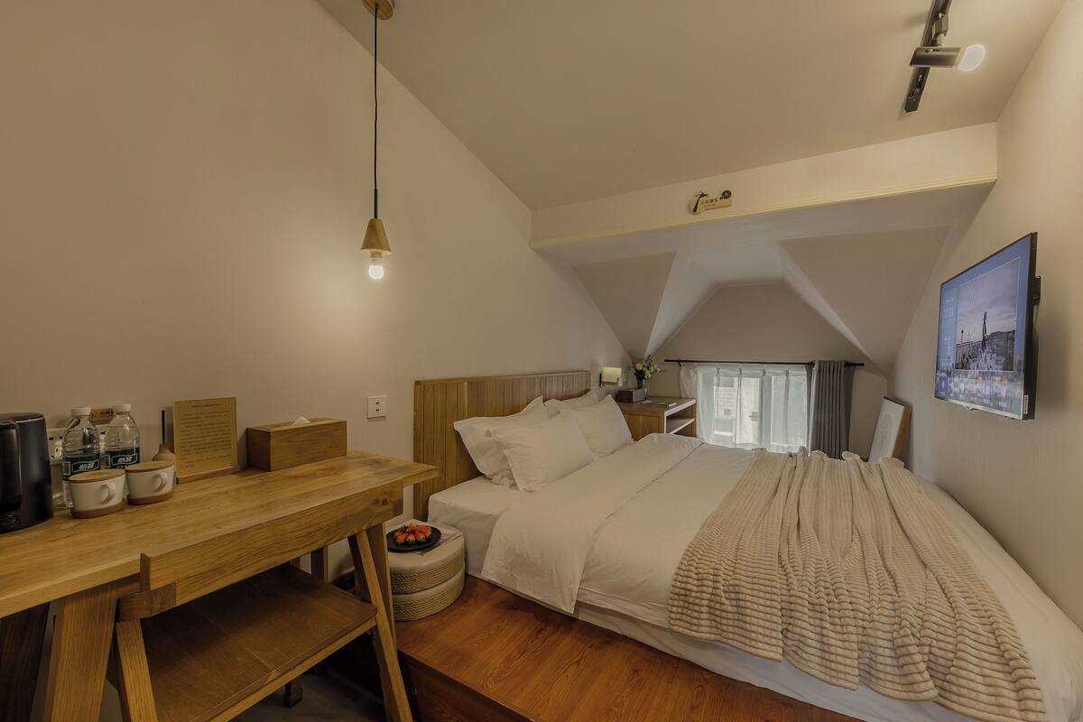 Ein Schlafzimmer mit einem hölzernen Schreibtisch, einem Bett mit einer beigefarbenen Decke, einer Deckenleuchte und einem gerahmten Bild an der Wand.