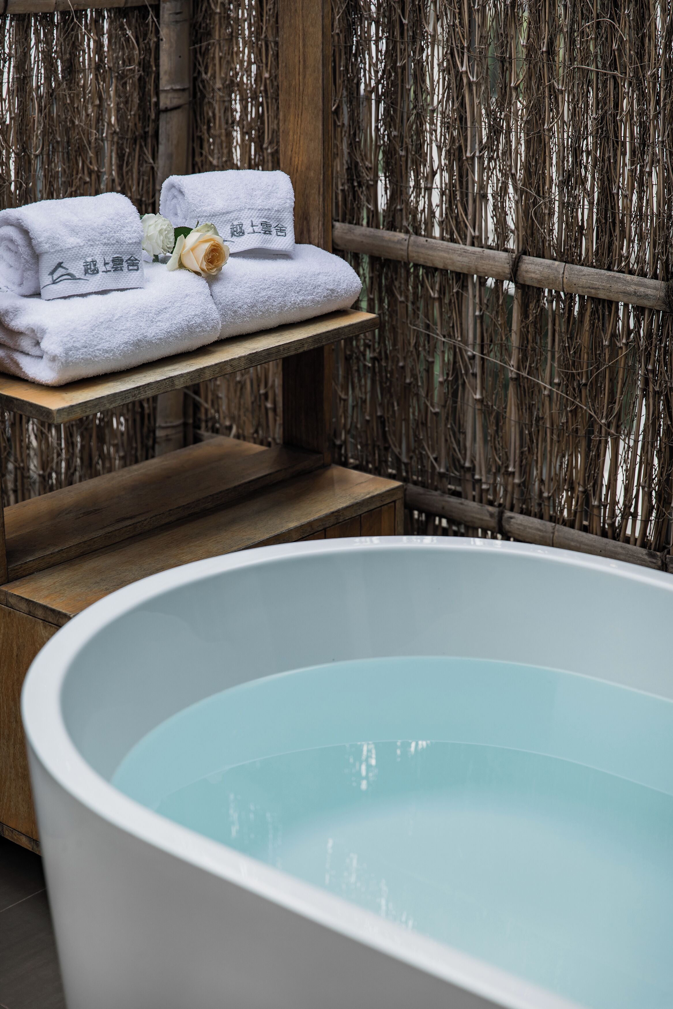 A spa area with a large bathtub, wooden shelves with towels, and a bamboo backdrop.