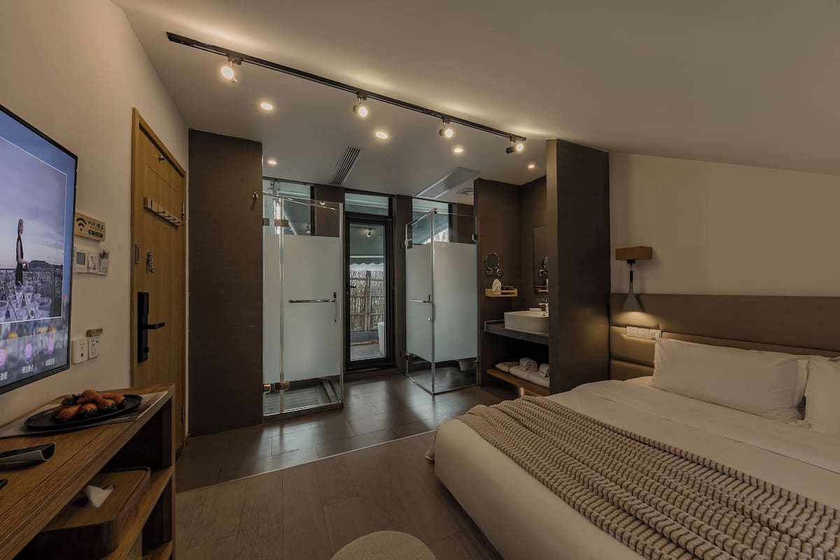 Ein modernes Hotelzimmer mit einem großen Bett, einem Fernseher auf einem Holztisch, einem Schreibtisch mit einem Laptop und einer Glastür, die auf einen Balkon führt.