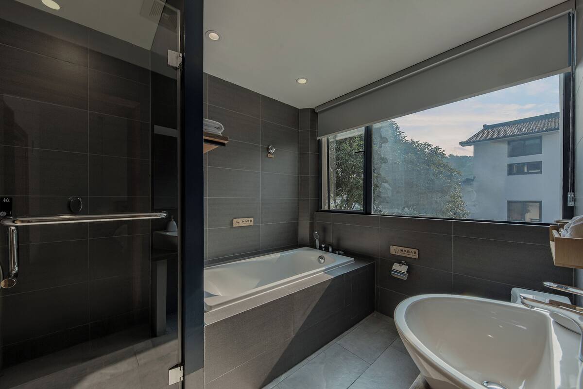 Ein modernes Badezimmer mit Duschkabine aus Glas, einer freistehenden Badewanne und einem großen Fenster mit Blick nach draußen.