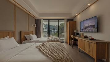 A hotel room with two beds, a wooden desk, a large window with a view of a house and trees, and a painting on the wall.