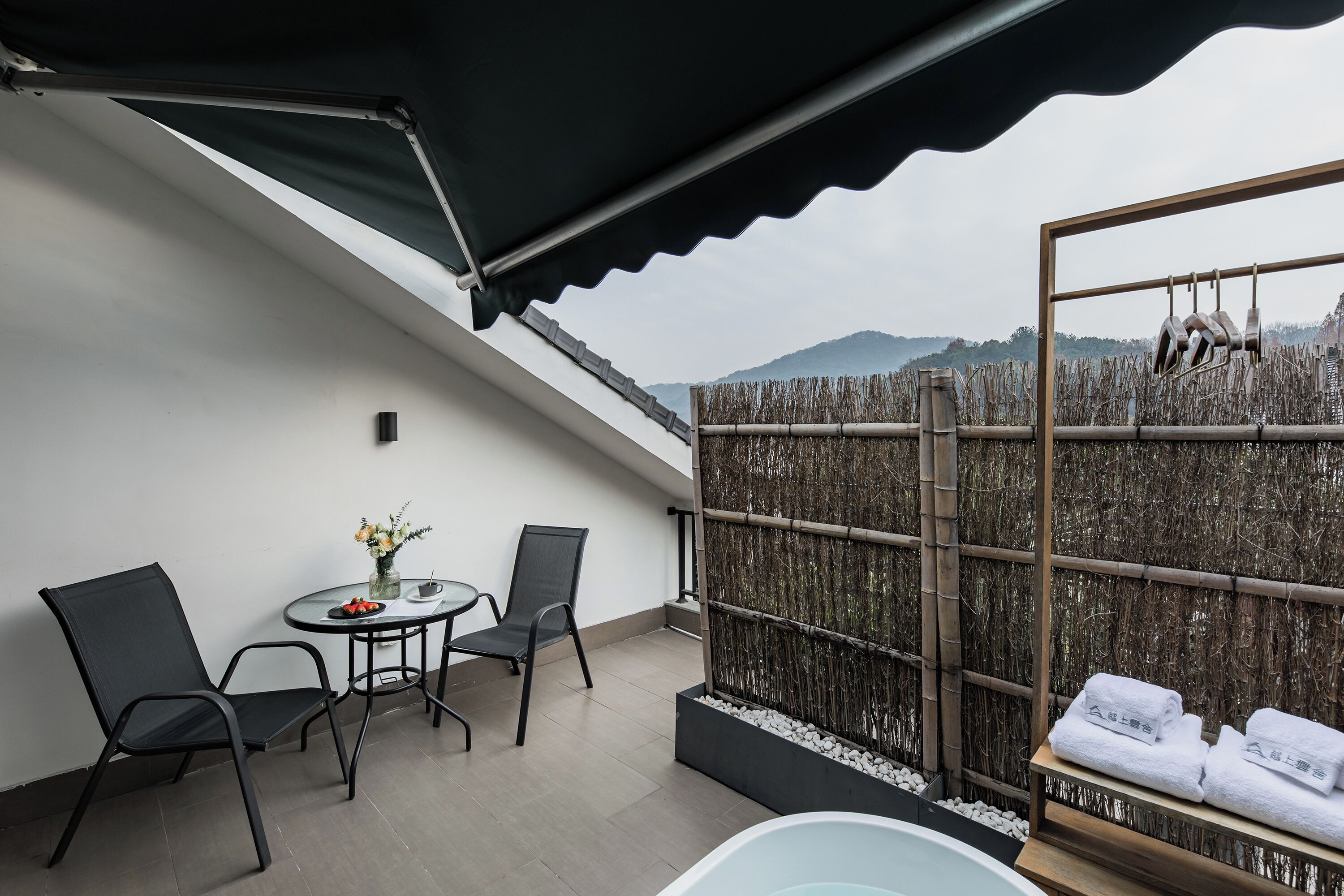 A balcony with a table, chairs, a small plant, a bamboo fence, and a shelf with towels.