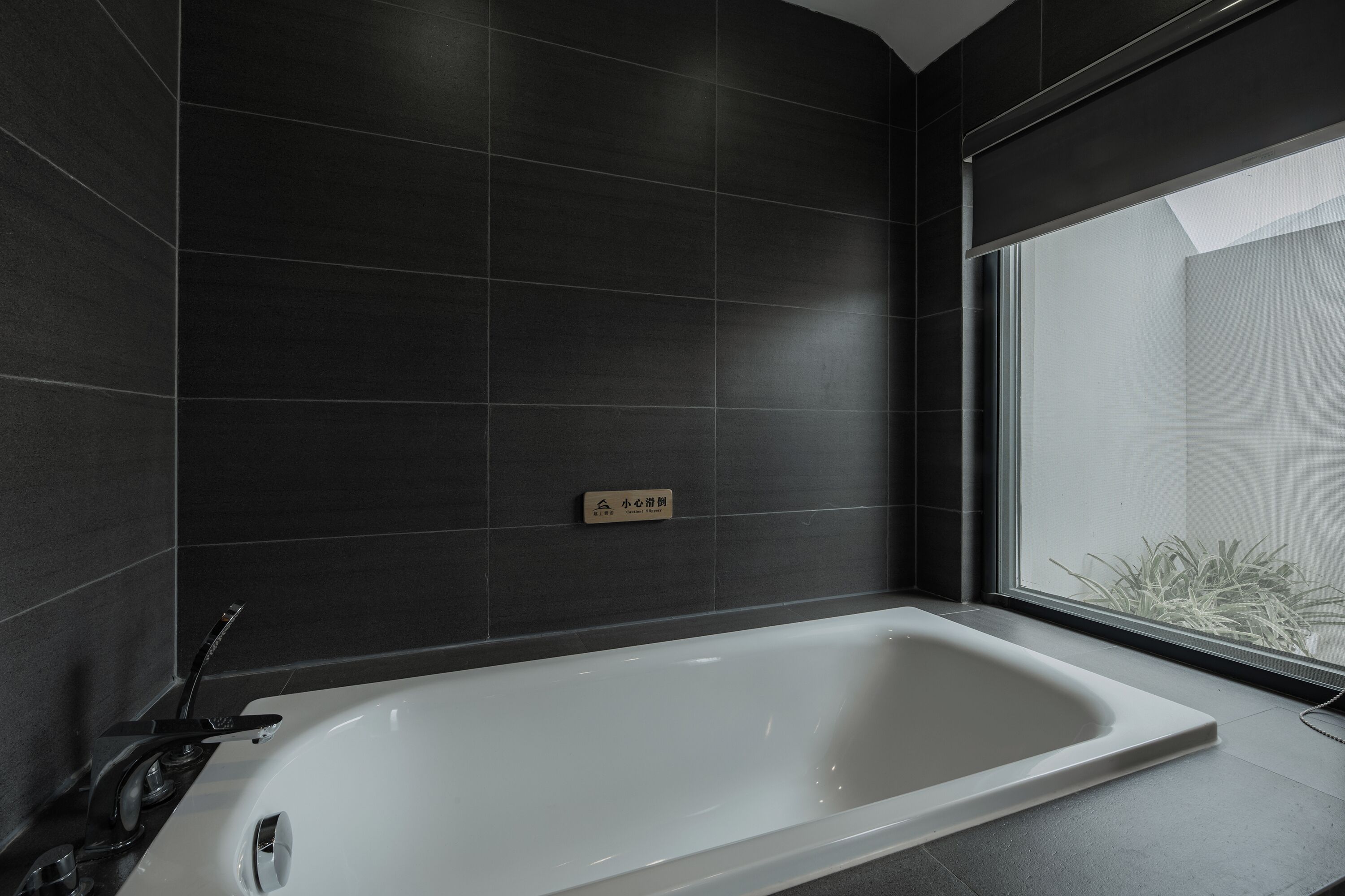A modern bathroom with a large bathtub, dark tiled walls, and a window providing a view of greenery outside.