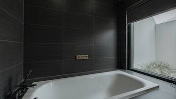 A modern bathroom with a large bathtub, dark tiled walls, and a window providing a view of greenery outside.