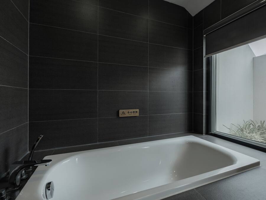 A modern bathroom with a large bathtub, dark tiled walls, and a window providing a view of greenery outside.