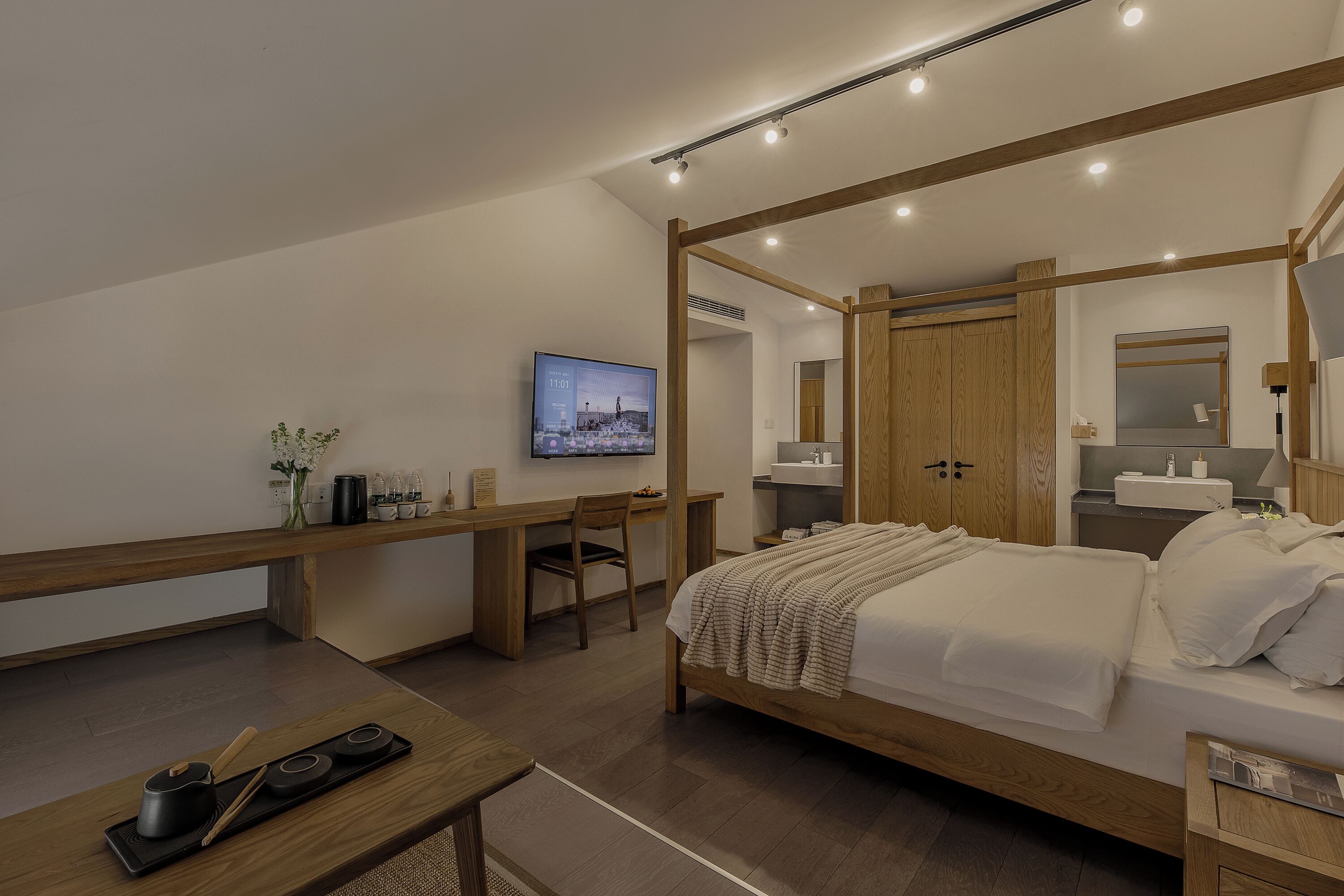 A modern bedroom with a wooden four-poster bed, a desk with a chair, a television, and a bathroom with a mirror and sink.