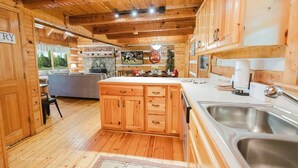 A wooden cabin interior with a kitchen area featuring wooden cabinets, a central island, and a large window overlooking a scenic outdoor area.