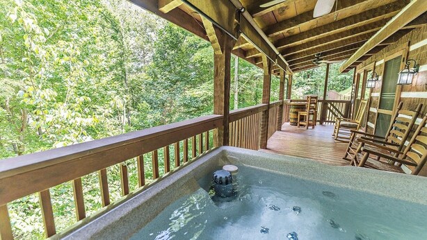 A wooden deck with a hot tub, wooden railings, and a view of a forest.
