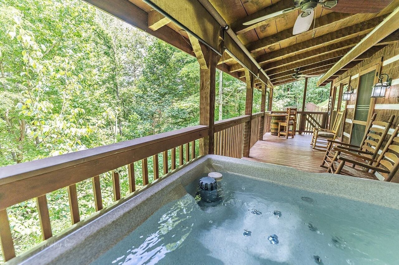 A wooden deck with a hot tub, surrounded by wooden railings and chairs, overlooking a forested area.