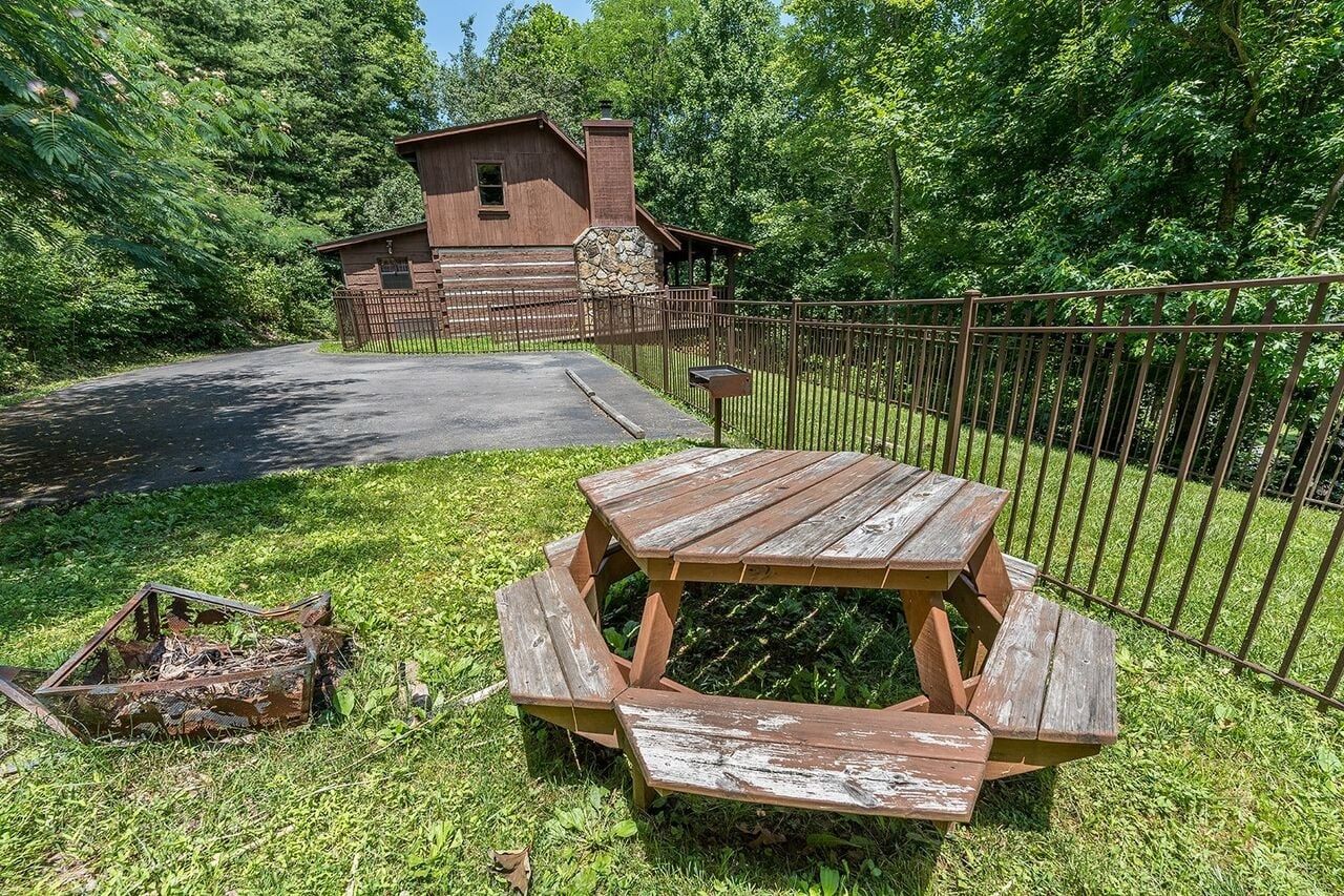 A rustic wooden cabin with a stone chimney, surrounded by a metal fence, and a picnic table in the foreground.