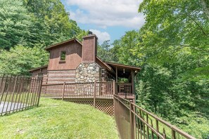 Un chalet rustique avec une façade en pierre, une terrasse en bois et une cour clôturée, le tout entouré d’arbres.