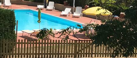 Piscina con sillas de descanso y sombrillas, rodeada por una cerca de madera y vegetación.
