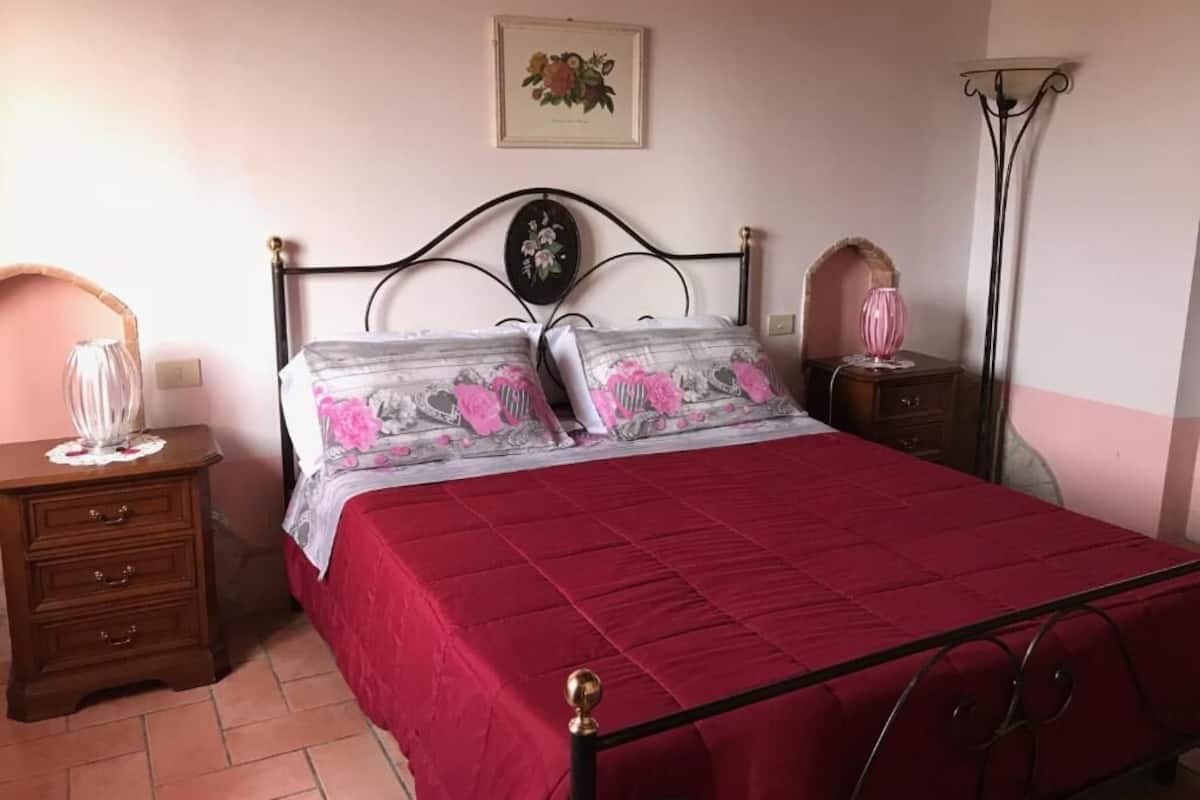 A bedroom with a metal bed frame, a red bedspread, two nightstands with lamps, and a wall-mounted picture.
