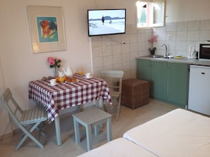 A small, cozy room with a checkered table set for breakfast, a television displaying a snowy landscape, a kitchenette with a sink and microwave, and a bed with a white sheet. - Harbour Inn Corfu (Corfu)