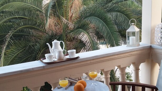 A balcony with a table set for two, featuring a pitcher, cups, and glasses of orange juice, with a view of palm trees.