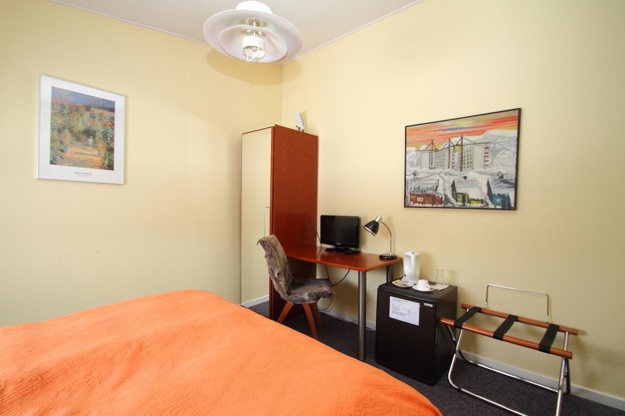Habitación de hotel con cama, escritorio y silla, televisor, una mesita con hervidor y radiador.
