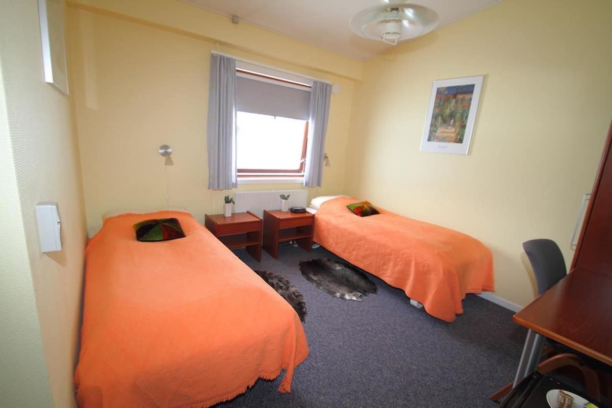 Une chambre avec deux lits, un bureau, une chaise, une fenêtre avec des rideaux et un tableau au mur.