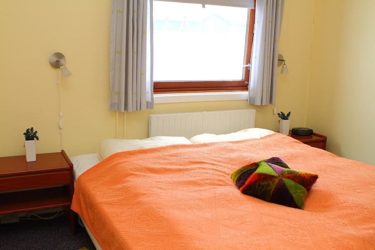 Une chambre à coucher avec un lit, une table de chevet, une lampe, une fenêtre avec des rideaux et un radiateur.