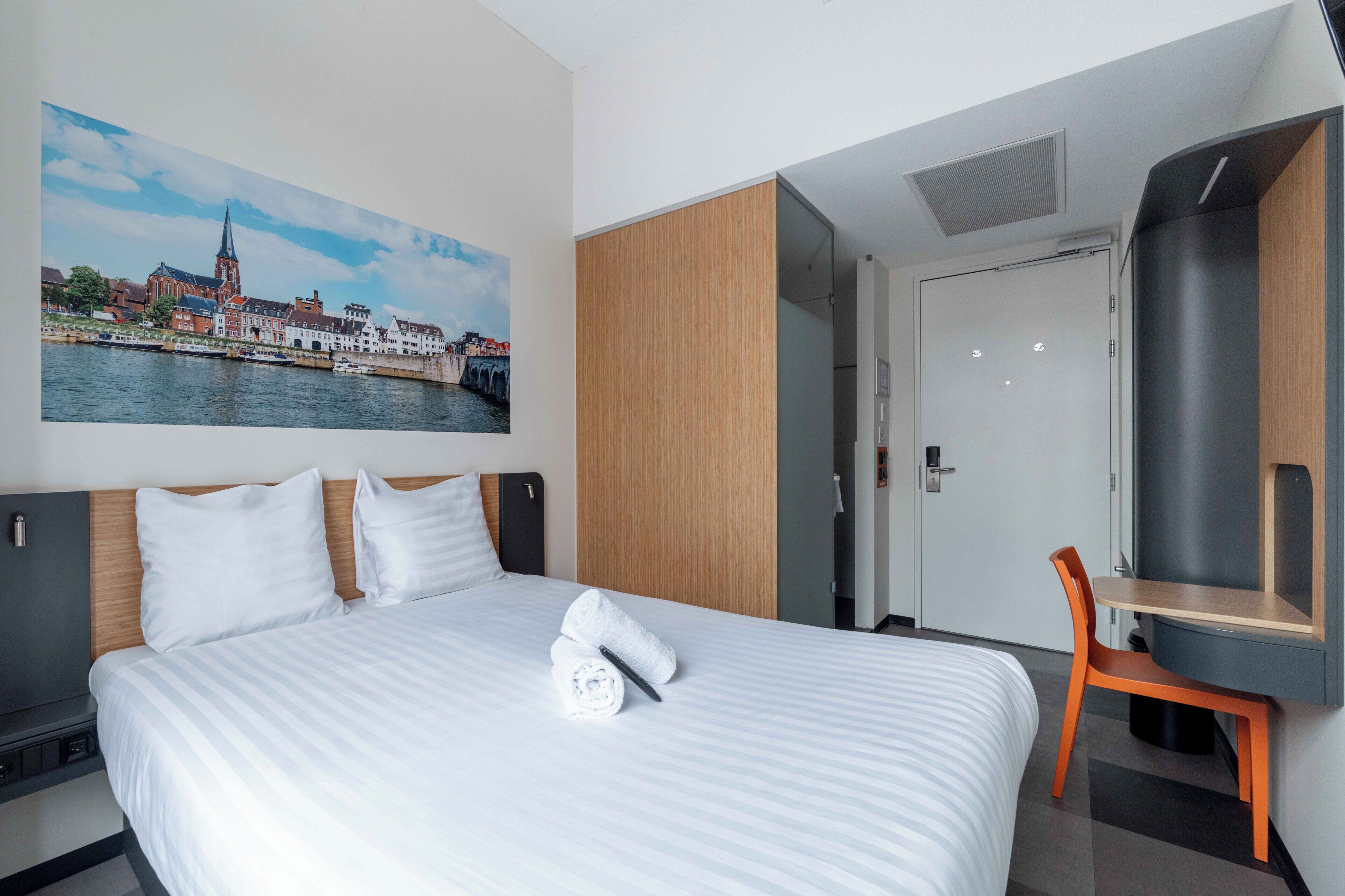 A hotel room with a bed, a desk, a chair, a painting of a cityscape, and a wardrobe.