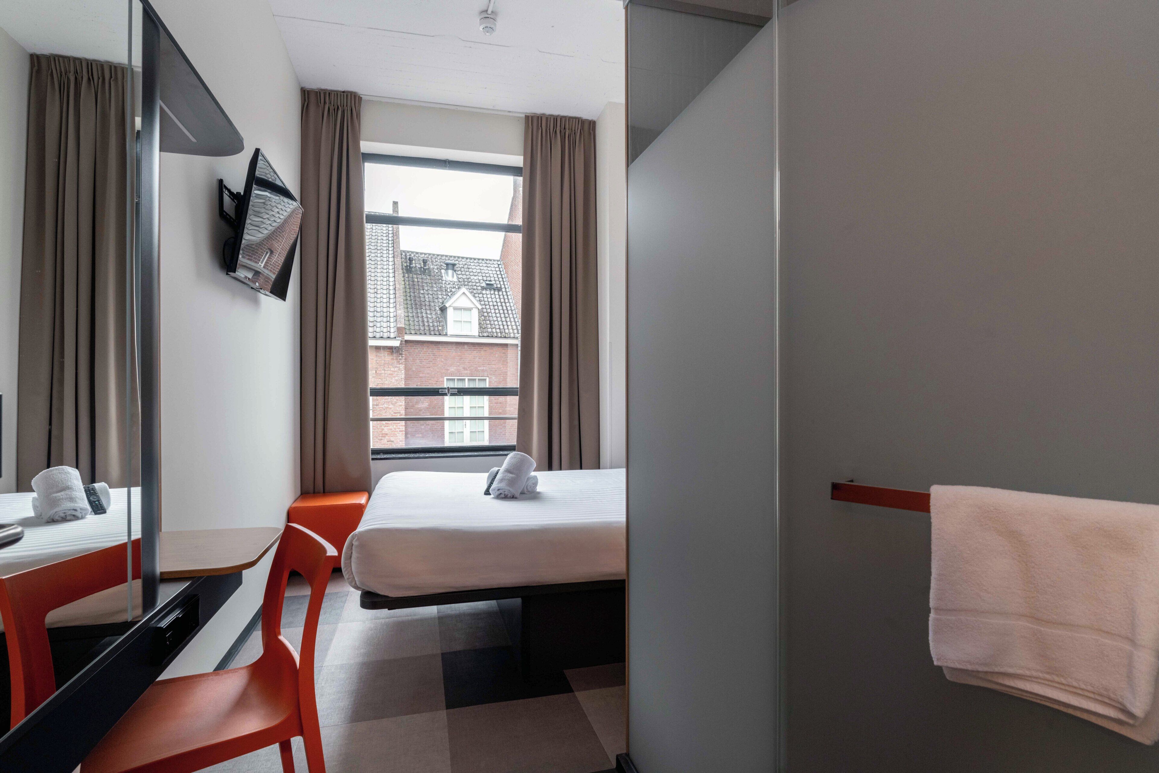 Une chambre d’hôtel équipée d’un lit, d’un bureau, d’une chaise, d’un téléviseur fixé au mur et d’une fenêtre avec des rideaux.