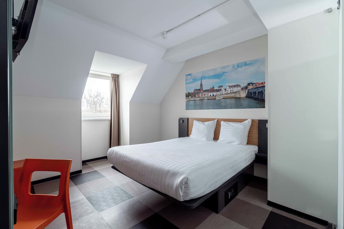 Ein modernes Hotelzimmer mit einem Bett, einem Fenster, einem Bild einer Brücke und einem roten Sessel.