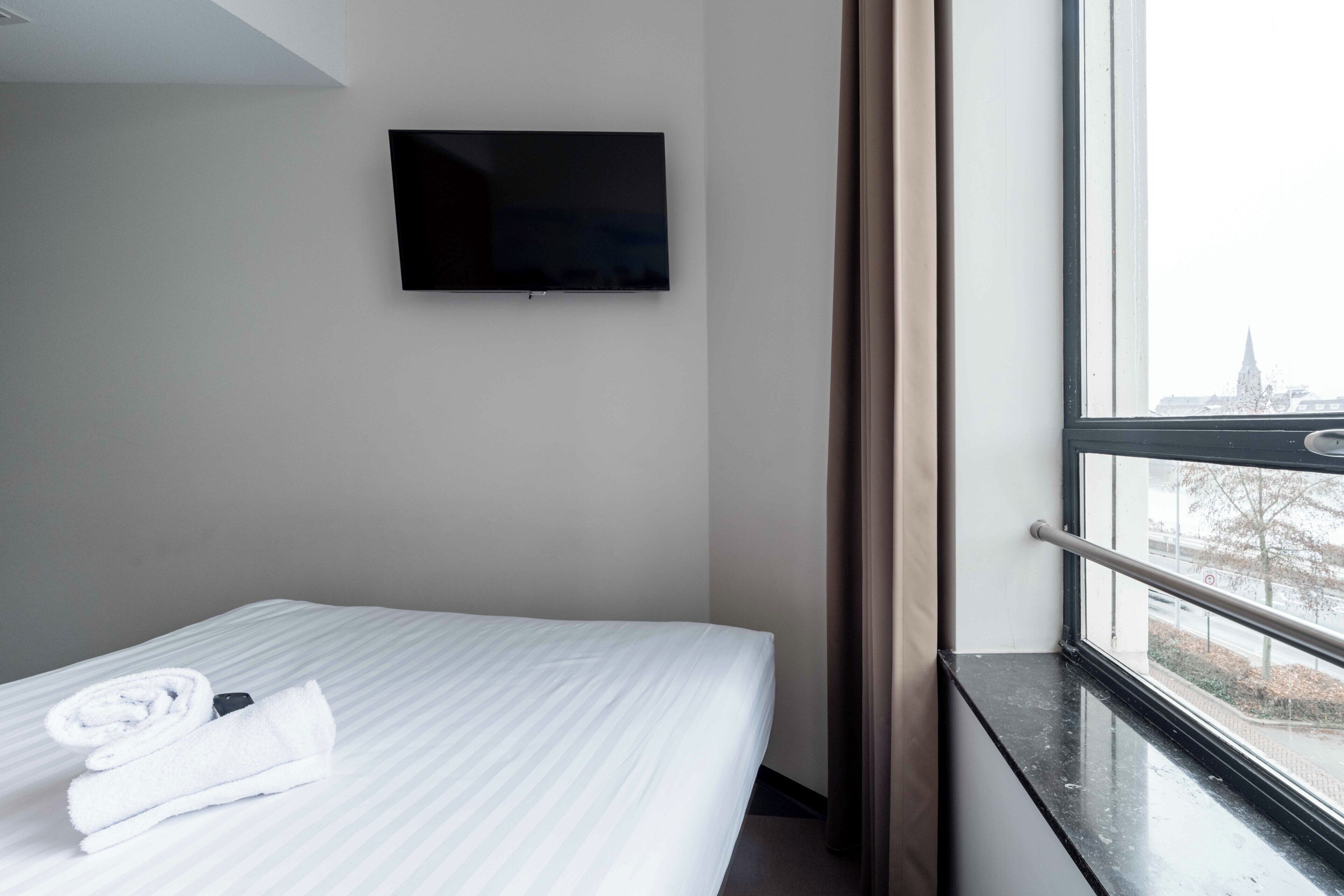 Une chambre d’hôtel avec un lit, un téléviseur fixé au mur et une fenêtre donnant sur un paysage urbain.