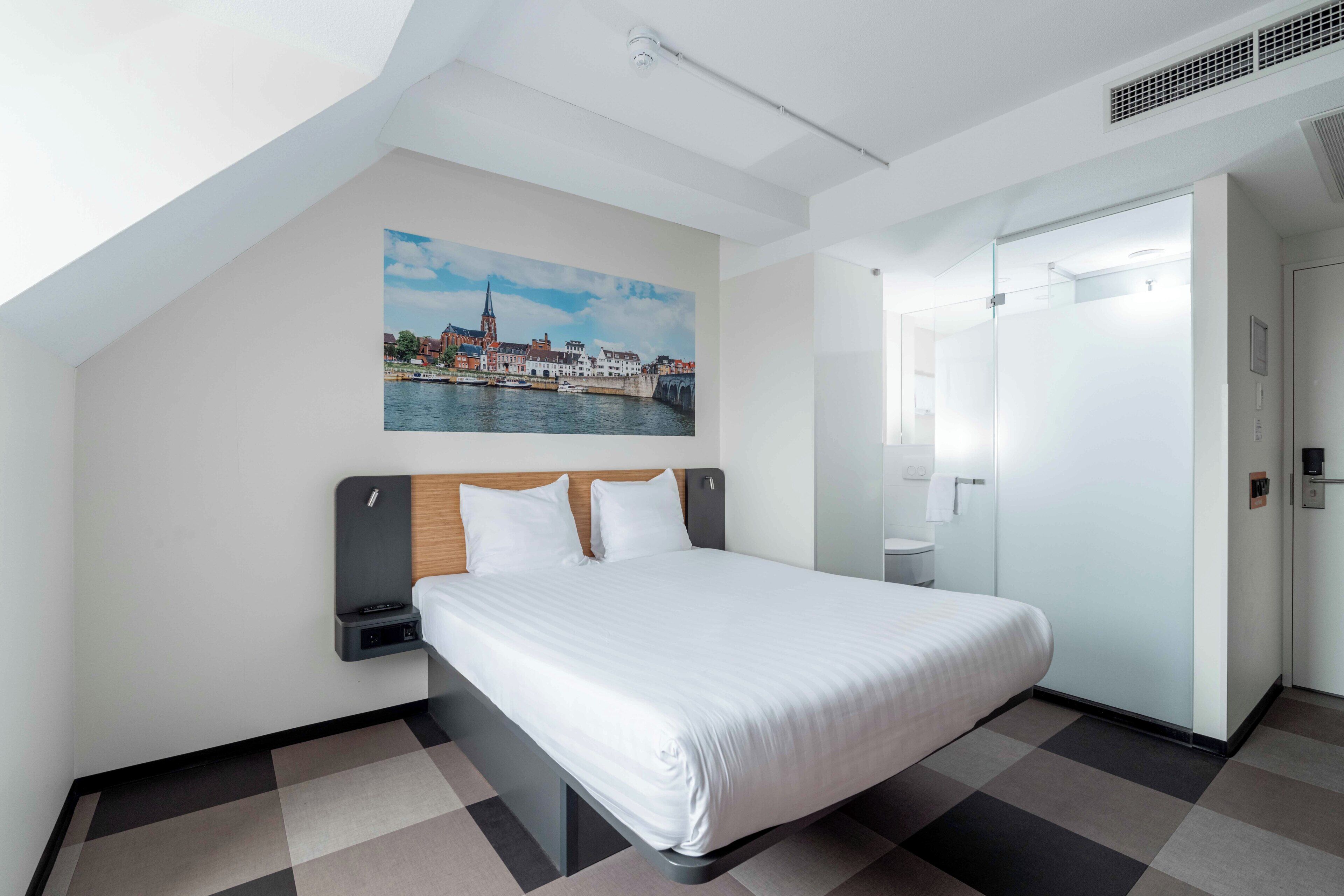 Une chambre d’hôtel moderne comprenant un lit, une table de chevet, un tableau accroché au mur, une salle de bain avec des toilettes et un lavabo, et une porte donnant sur une autre pièce.