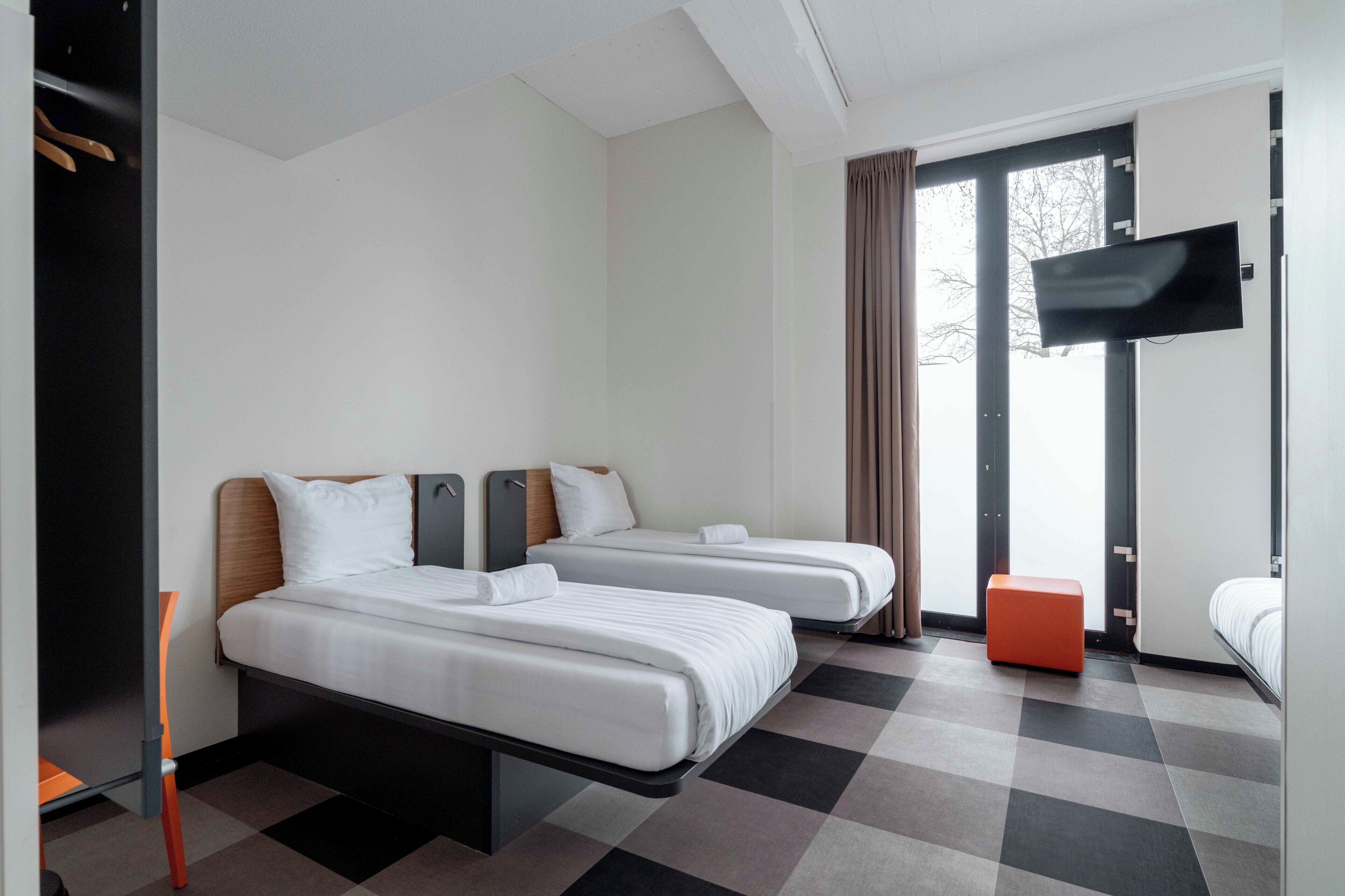 A hotel room with two beds, a flat-screen TV, a window with curtains, and a checkered floor.