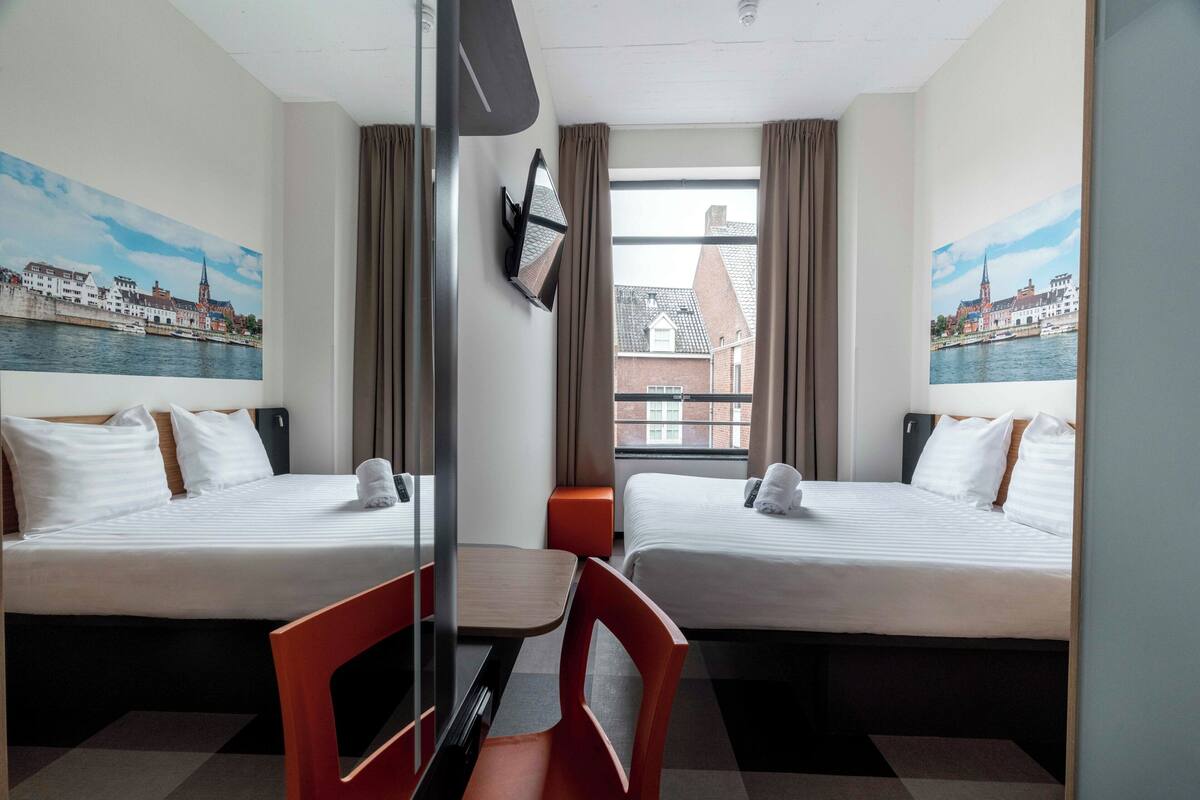 Ein Hotelzimmer mit zwei Betten, einem Schreibtisch, einem Sessel, einem Fernseher und einem Fenster mit Blick auf Gebäude und Wasser.