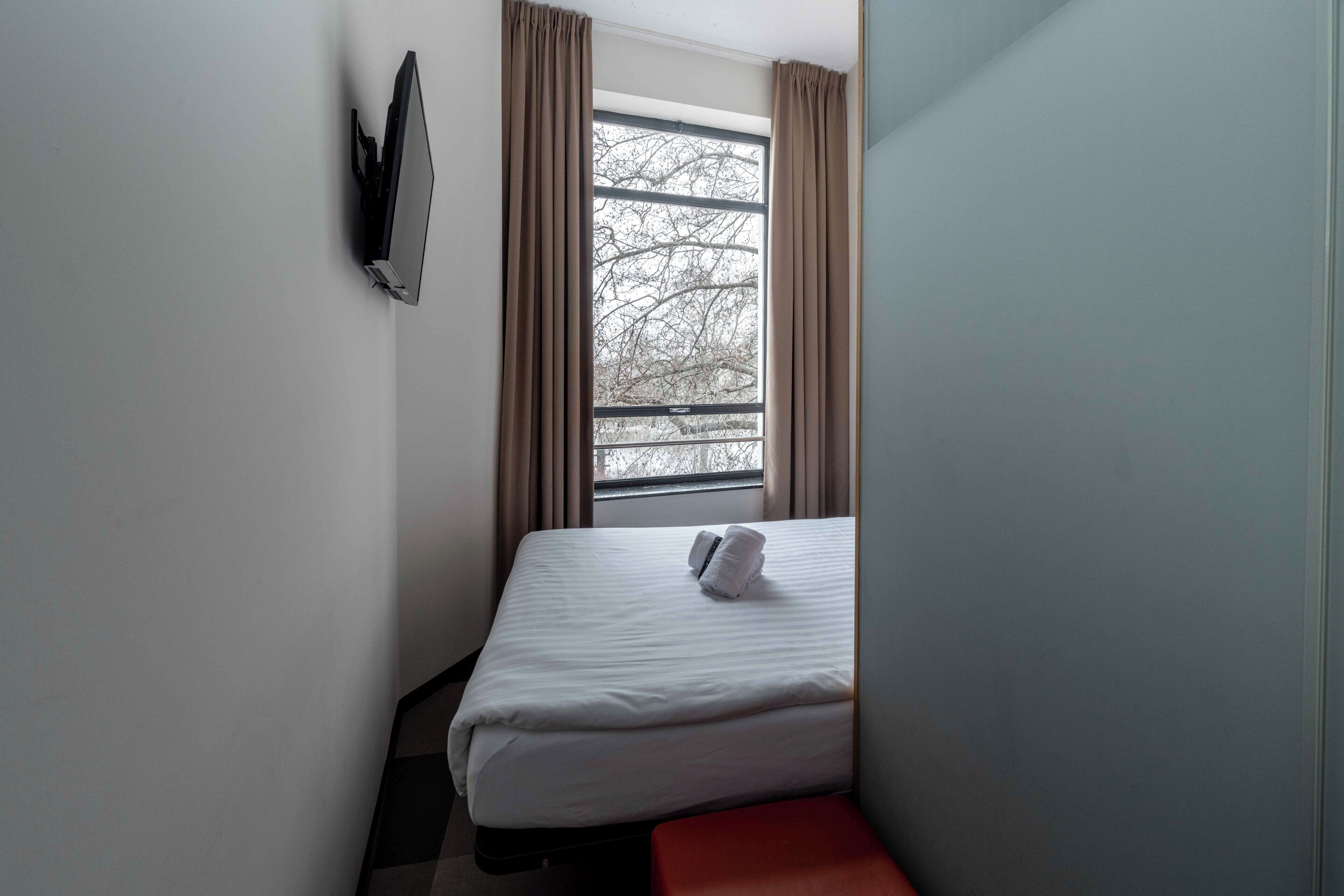 A small, single-bed room with a window, a flat-screen TV mounted on the wall, and a red bench.