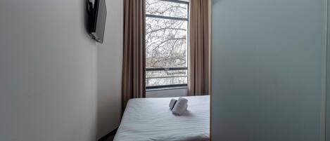 A small, single-bed room with a window, a flat-screen TV mounted on the wall, and a red bench.