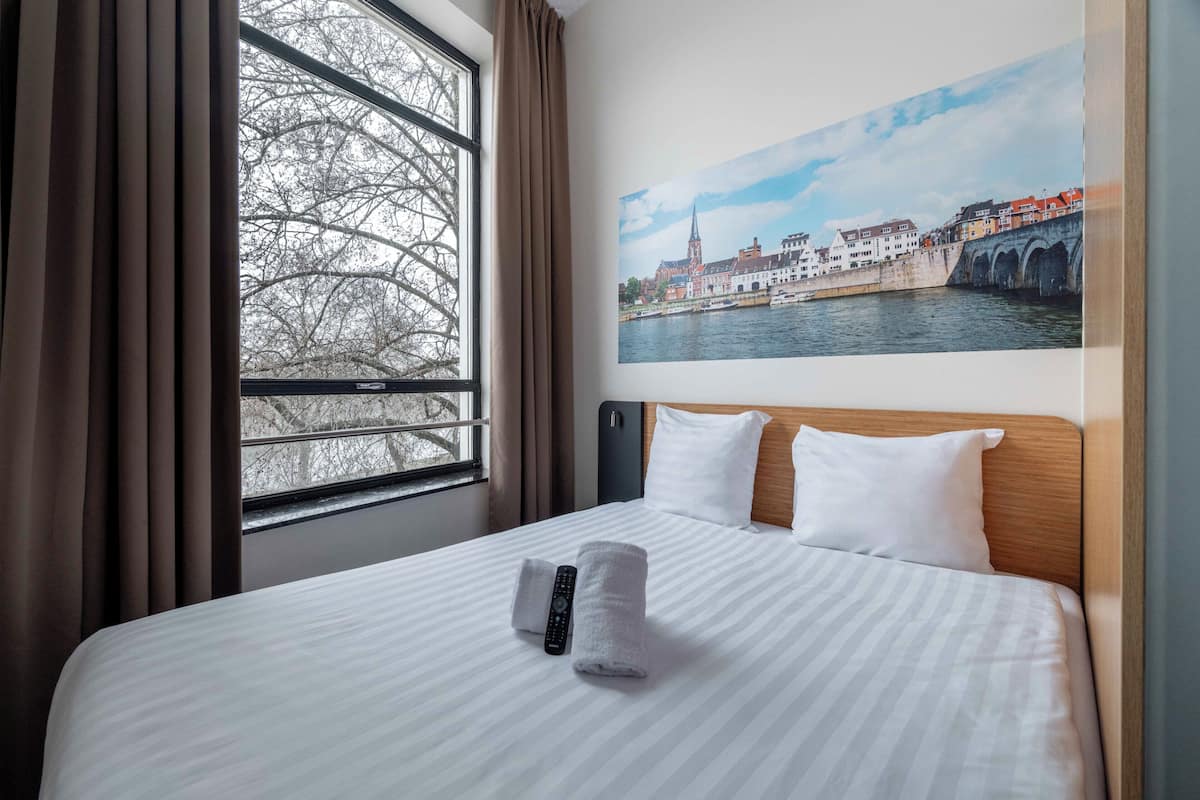 Ein Hotelzimmer mit einem großen Bett, einem Fenster mit Vorhängen, einem Stadtbild als Gemälde und einer Fernbedienung auf dem Bett.