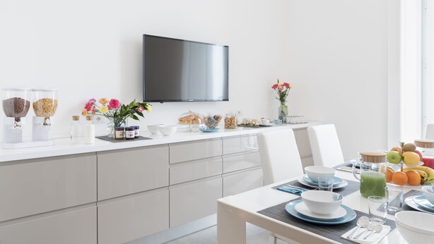 A modern kitchen with a flat-screen TV mounted above the counter, white cabinets, and a dining area with a table set for breakfast. - Lux B&B (Naples)