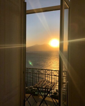 Un balcón con vistas a la puesta de sol sobre el mar, barandilla metálica y banco de madera.