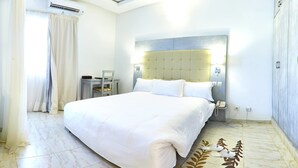 A modern hotel room with a large bed, a desk, and a chair. There is an air conditioning unit on the wall, a telephone, and a wardrobe.