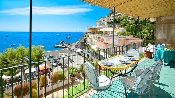 A balcony with a table and chairs overlooking a coastal town with buildings and boats.