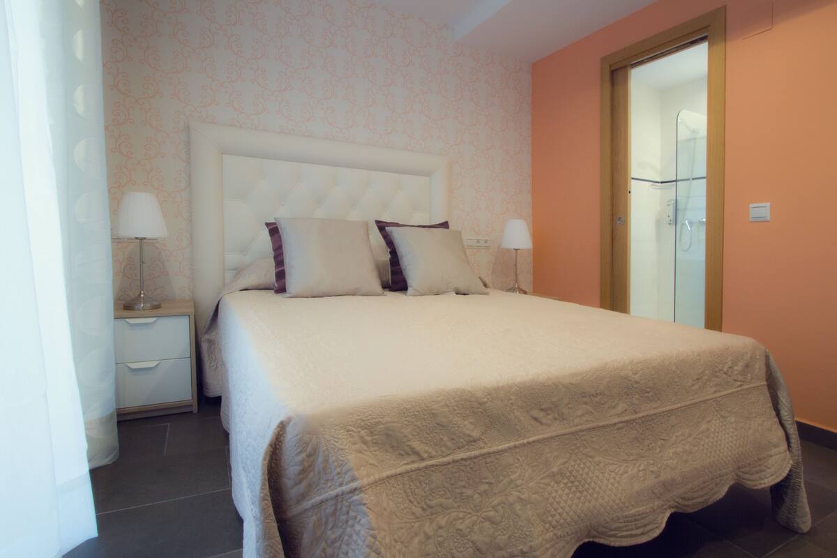 A neatly made bed with a headboard, two bedside tables with lamps, and a door to a bathroom.