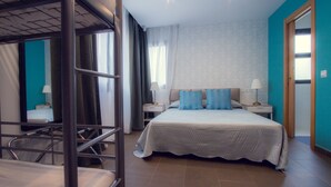 Une chambre d’hôtel avec un lit superposé, un lit simple avec des oreillers bleus, deux tables de chevet avec des lampes et une grande fenêtre avec des rideaux.