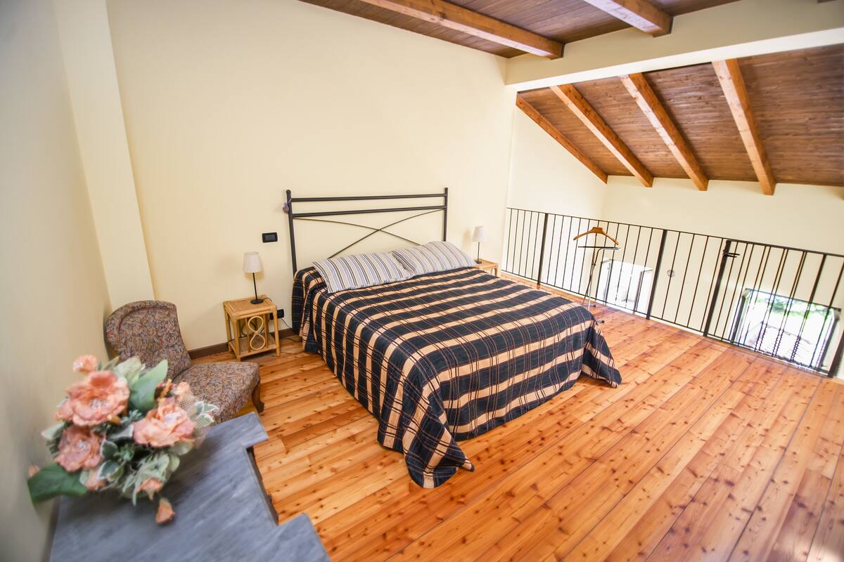 A bedroom with a wooden floor, a bed with a striped bedspread, a chair, a nightstand with a lamp, and a balcony with a railing.